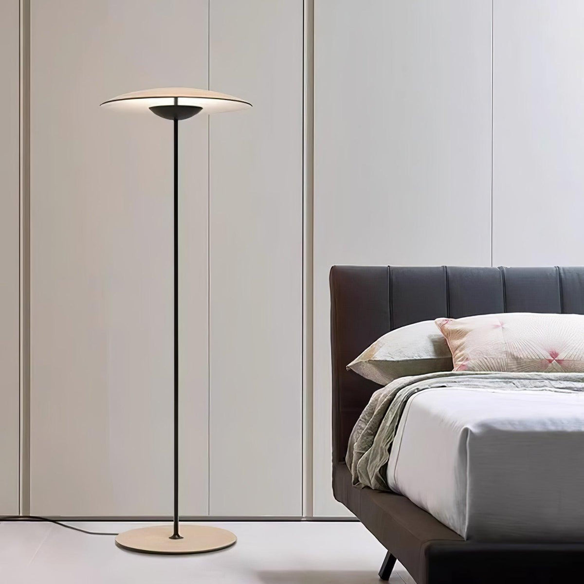 A Clérachic floor lamp stands beside a bed with a dark ribbed headboard, in front of tall pale wardrobe doors.