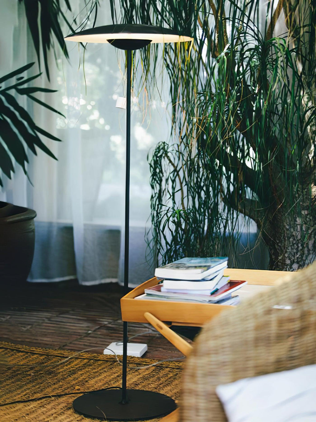 A matte black Clérachic floor lamp stands among tall plants and sheer curtains, beside a wicker chair and a tray of books.