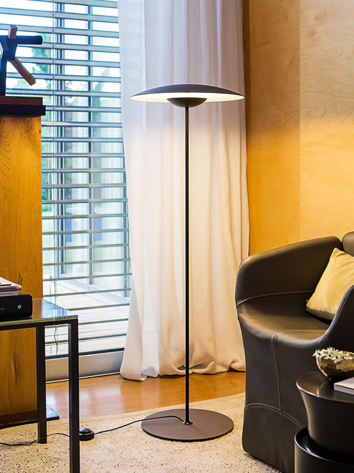 A matte black Clérachic floor lamp stands by a white curtain and a window with slatted blinds, near a dark leather chair.