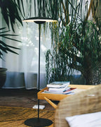 Tighter crop of a matte black Clérachic floor lamp among leafy plants, with a wicker chair and a stack of books beside it.