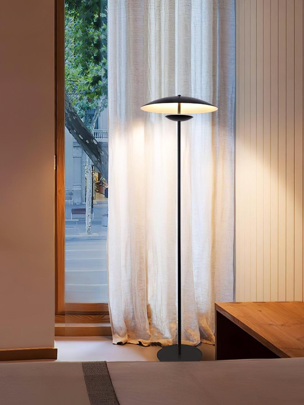 A matte black Clérachic floor lamp stands before a floor-length sheer curtain and a glass door onto the street.