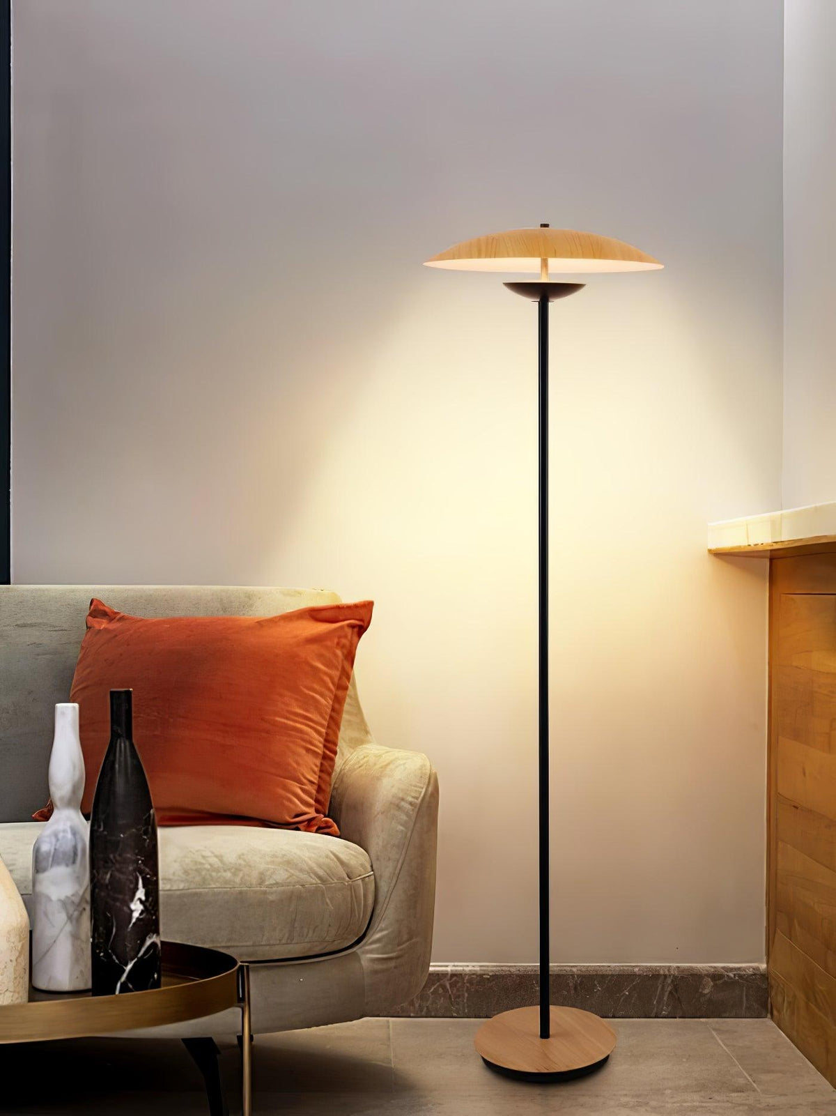 A Clérachic floor lamp casts a warm glow over a cream armchair with an orange cushion and marble vases on a side table.