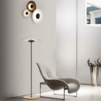 A Clérachic floor lamp stands in a pale room beside a grey lounge chair, with a trio of round wall lights above.