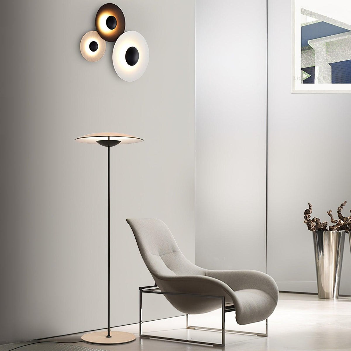 A Clérachic floor lamp stands in a pale room beside a grey lounge chair, with a trio of round wall lights above.