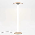A Clérachic floor lamp on a soft cream backdrop, its glowing disc shade held on a thin black stem above a wood-grain base.