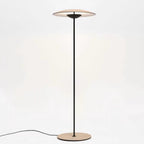 A Clérachic floor lamp on a soft cream backdrop, its glowing disc shade held on a thin black stem above a wood-grain base.