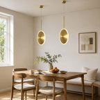 Two vertical Clérachic disc pendants in brushed gold hang over a wooden dining table in a bright corner with a window and a framed print.