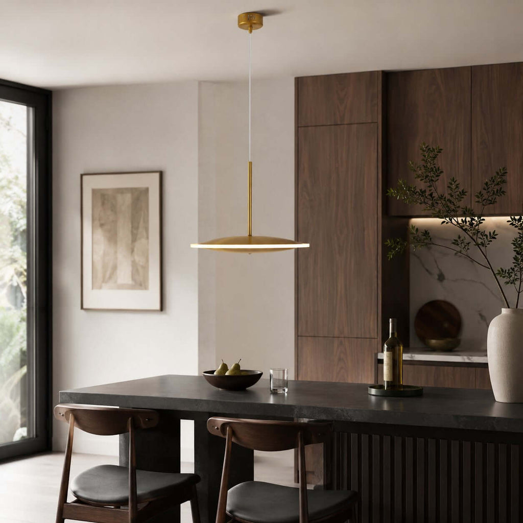 A slim horizontal Clérachic disc pendant in brushed gold hangs above a dark table, with walnut cabinets and a tall window behind.