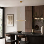 A slim horizontal Clérachic disc pendant in brushed gold hangs above a dark table, with walnut cabinets and a tall window behind.