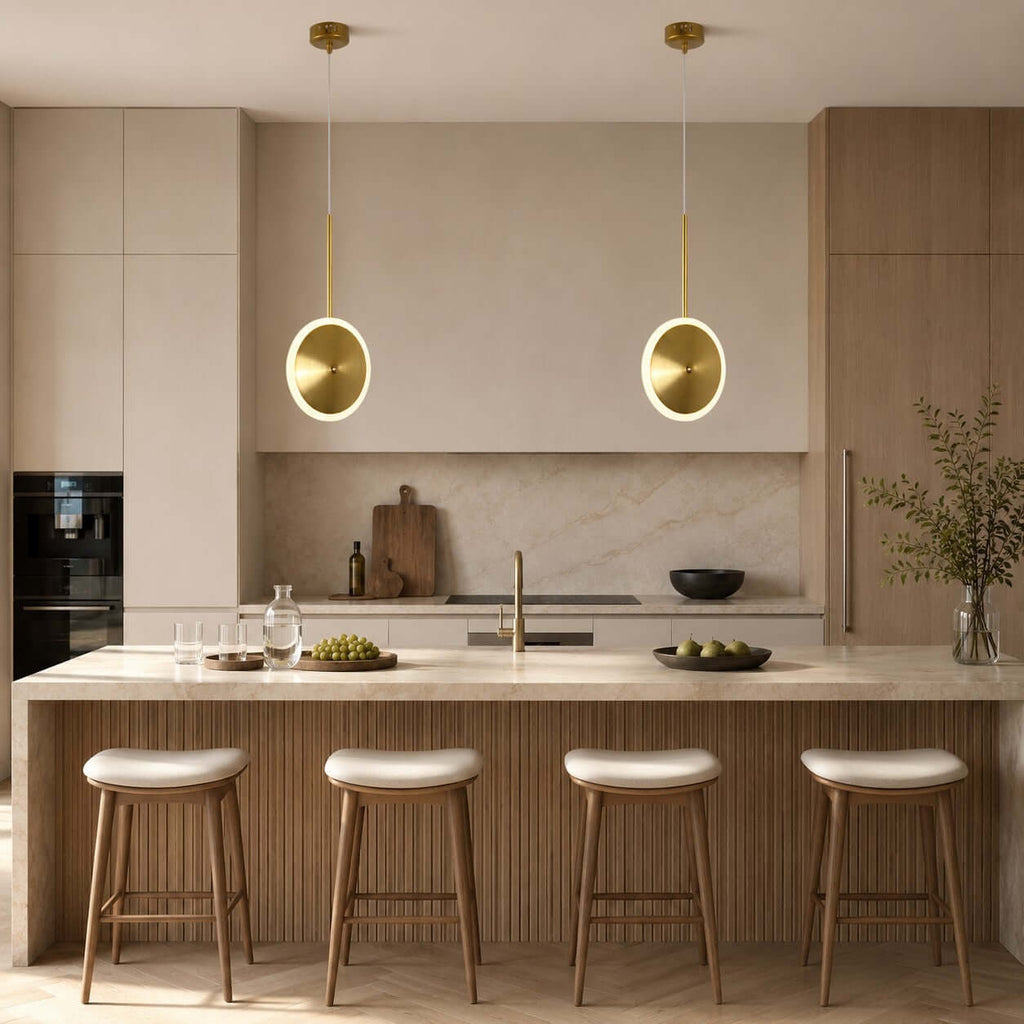 Two vertical Clérachic disc pendants in brushed gold hang over a pale stone kitchen island with four cream-topped wooden stools.