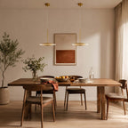 Two horizontal Clérachic disc pendants in brushed gold hang over a wooden dining table with a framed rust-coloured print and a plant behind.
