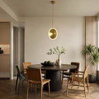 One vertical Clérachic disc pendant in brushed gold hangs over a round dark dining table with a mix of wooden chairs and a large palm.