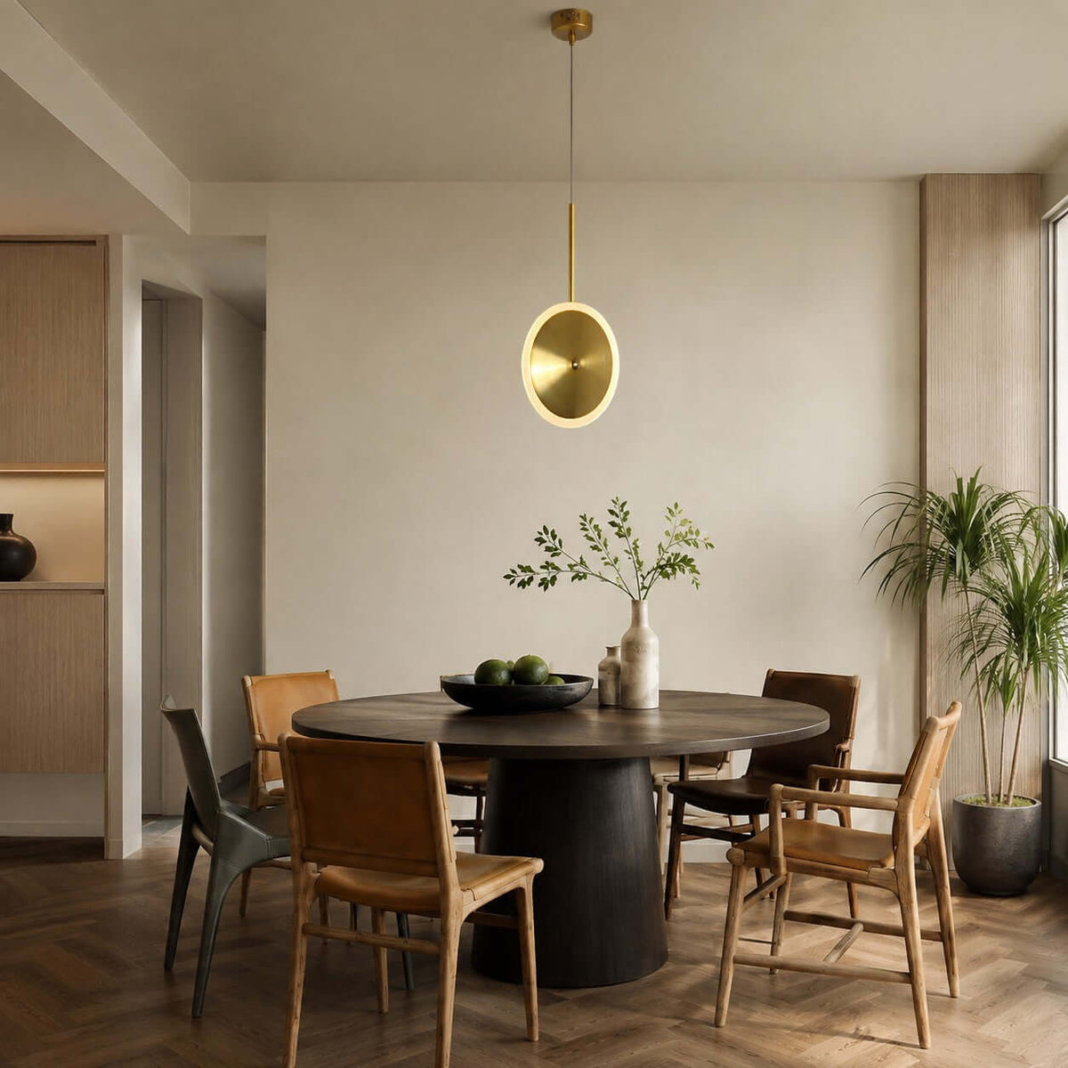 One vertical Clérachic disc pendant in brushed gold hangs over a round dark dining table with a mix of wooden chairs and a large palm.