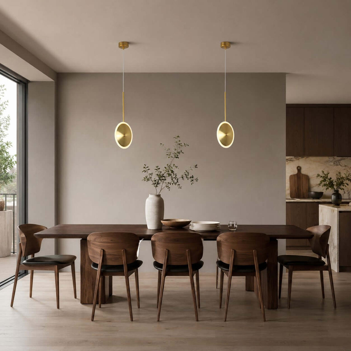 Two small vertical Clérachic disc pendants in brushed gold hang over a long dark dining table with leather chairs and an open kitchen behind.