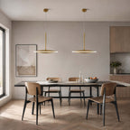 Two horizontal Clérachic disc pendants in brushed gold hang over a long dark table with tan leather chairs, a framed print and a tall window.