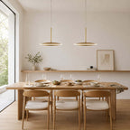 Two horizontal Clérachic disc pendants in brushed gold hang over a long oak dining table with white-seated wooden chairs and a floating shelf.