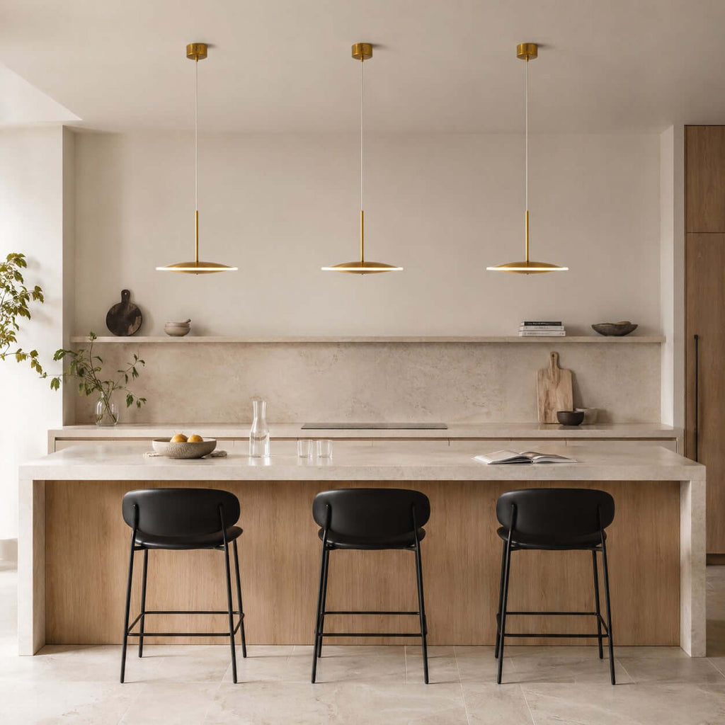 Three horizontal Clérachic disc pendants in brushed gold hang over a stone kitchen island with black-seated stools and a marble splashback.