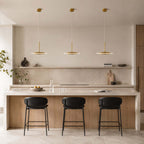 Three horizontal Clérachic disc pendants in brushed gold hang over a stone kitchen island with black-seated stools and a marble splashback.