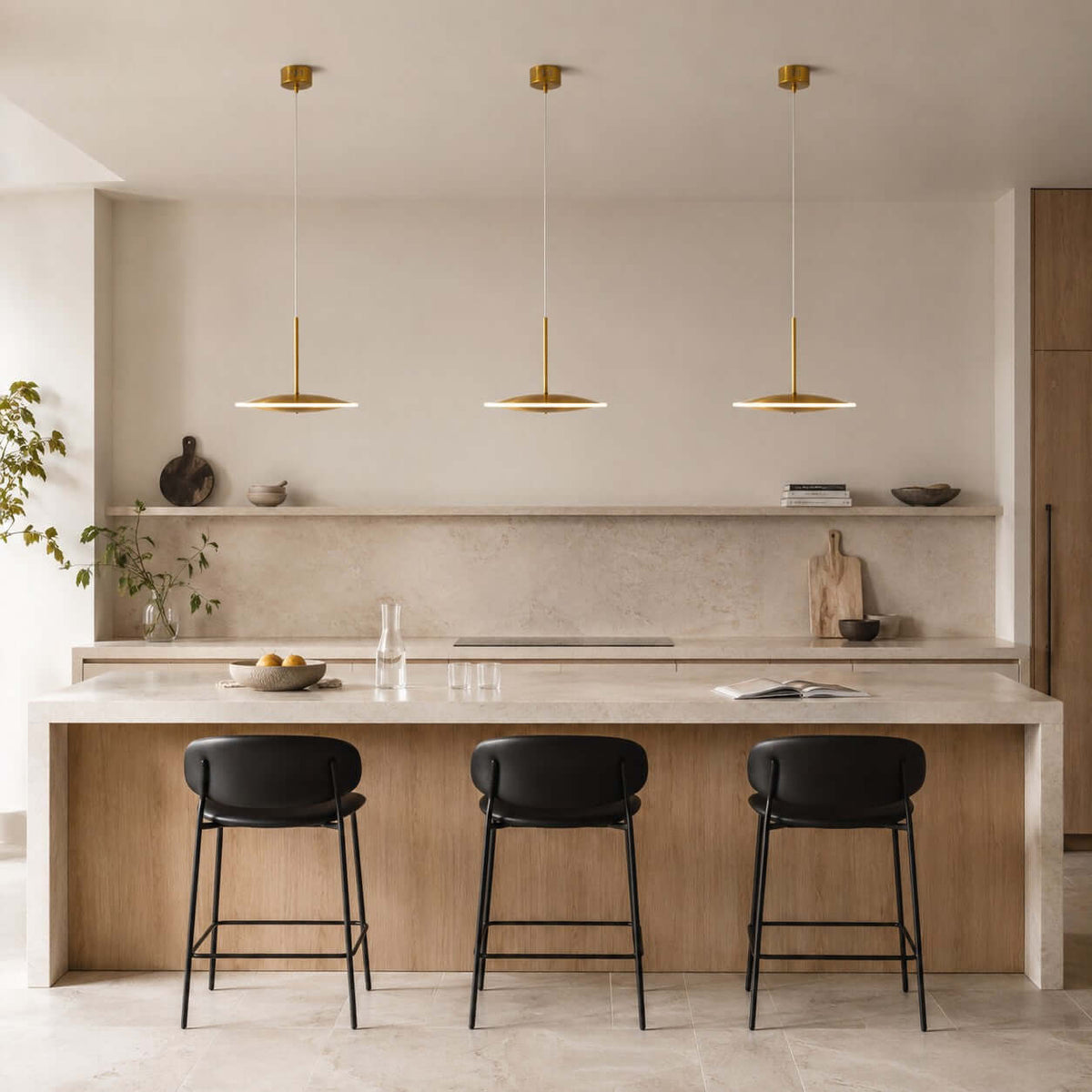 Three horizontal Clérachic disc pendants in brushed gold hang over a stone kitchen island with black-seated stools and a marble splashback.