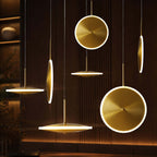 A cluster of Clérachic brushed gold disc pendants, horizontal and vertical, glow at different heights against a dark slatted wall.