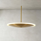 Close view of a horizontal Clérachic disc pendant in brushed gold, its soft glowing ring edge and slim stem against a pale plaster wall.