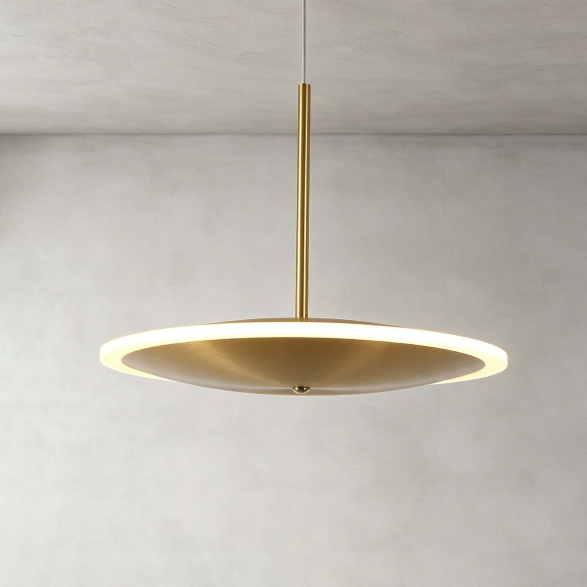 Close view of a horizontal Clérachic disc pendant in brushed gold, its soft glowing ring edge and slim stem against a pale plaster wall.