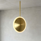 Close view of a vertical Clérachic disc pendant in brushed gold, its brushed round face and glowing frosted ring hung from a slim brass stem.