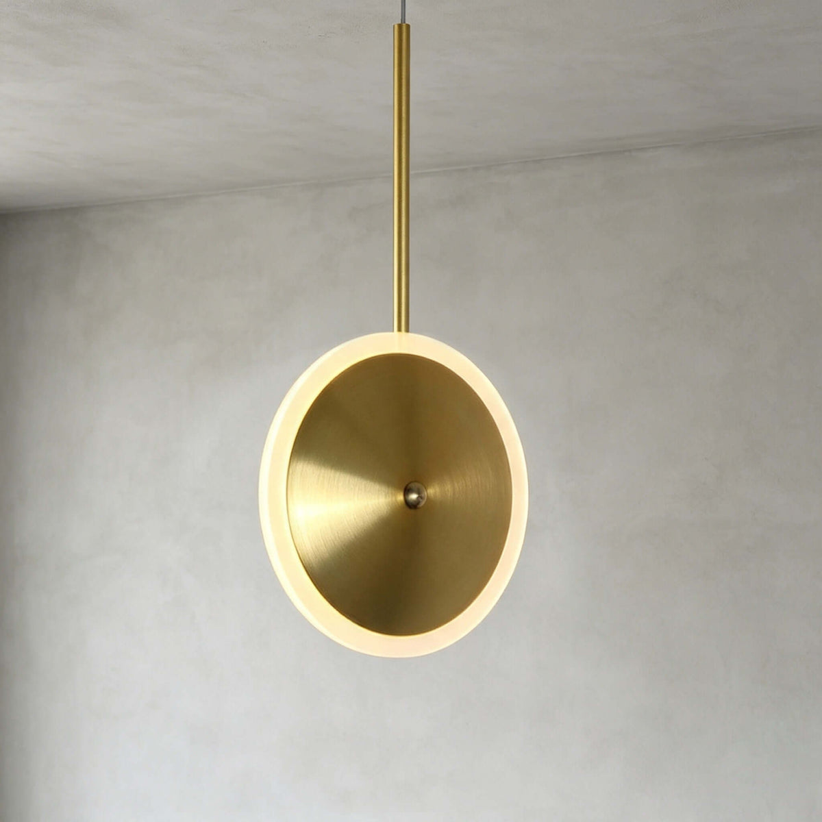 Close view of a vertical Clérachic disc pendant in brushed gold, its brushed round face and glowing frosted ring hung from a slim brass stem.