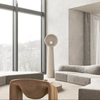 White textured Clérachic floor lamp beside tall windows in a calm living room with a low grey sofa and a rounded wooden chair.