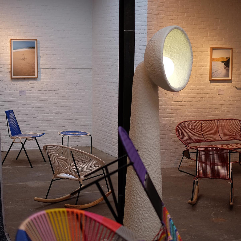 Clérachic floor lamp with a lit bowl shade turned toward the viewer, in a brick-walled room with bright woven chairs.