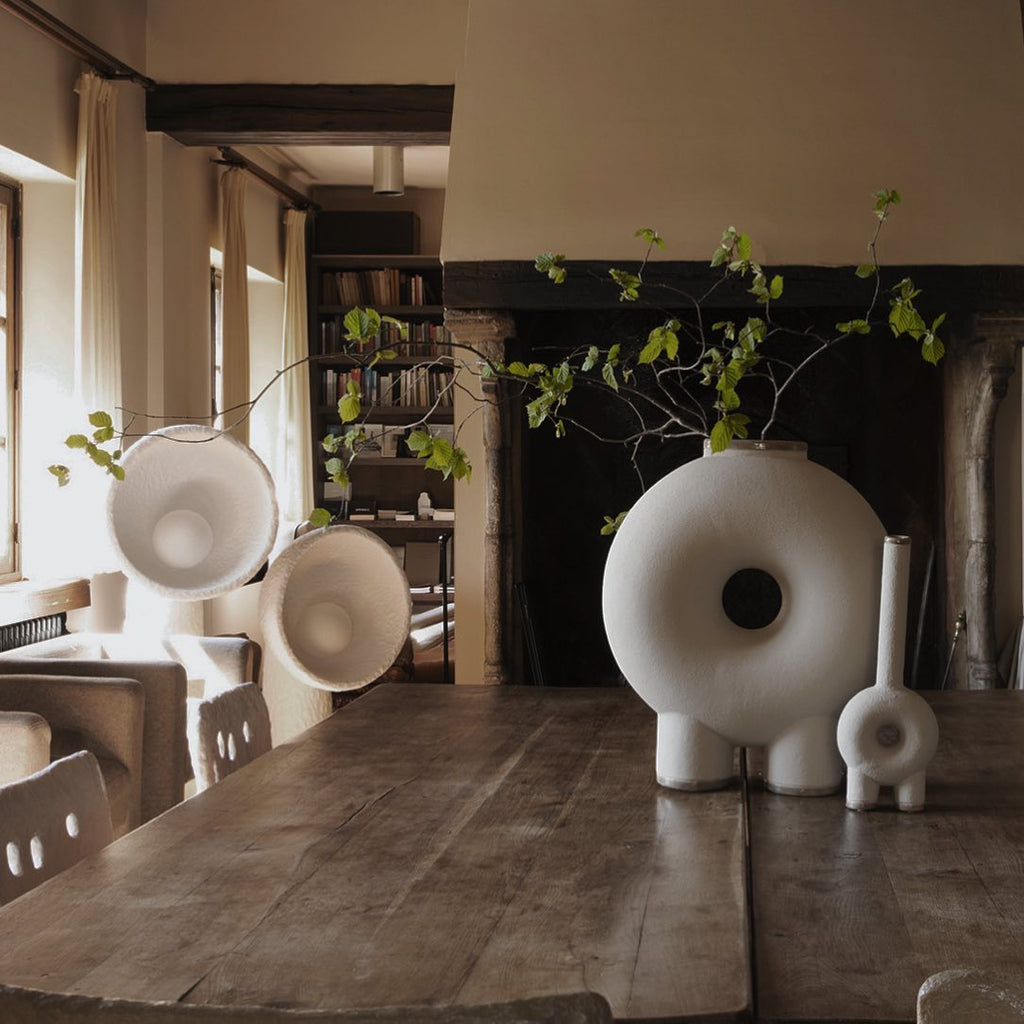 Two Clérachic lamps with bowl shades on a long wooden table in front of a fireplace, with a round white vase and a branch.
