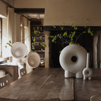 Two Clérachic lamps with bowl shades on a long wooden table in front of a fireplace, with a round white vase and a branch.