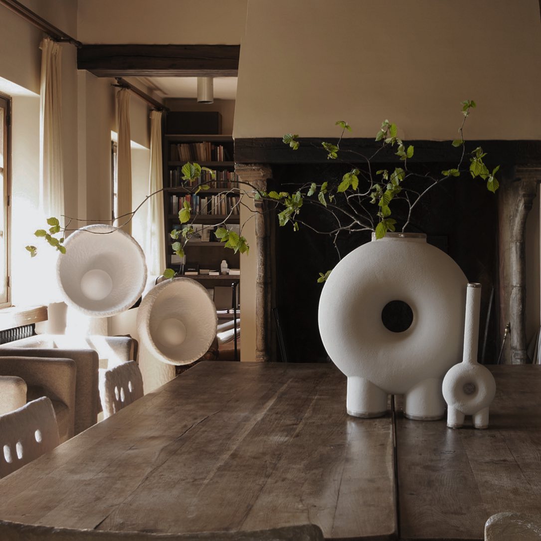 Two Clérachic lamps with bowl shades on a long wooden table in front of a fireplace, with a round white vase and a branch.