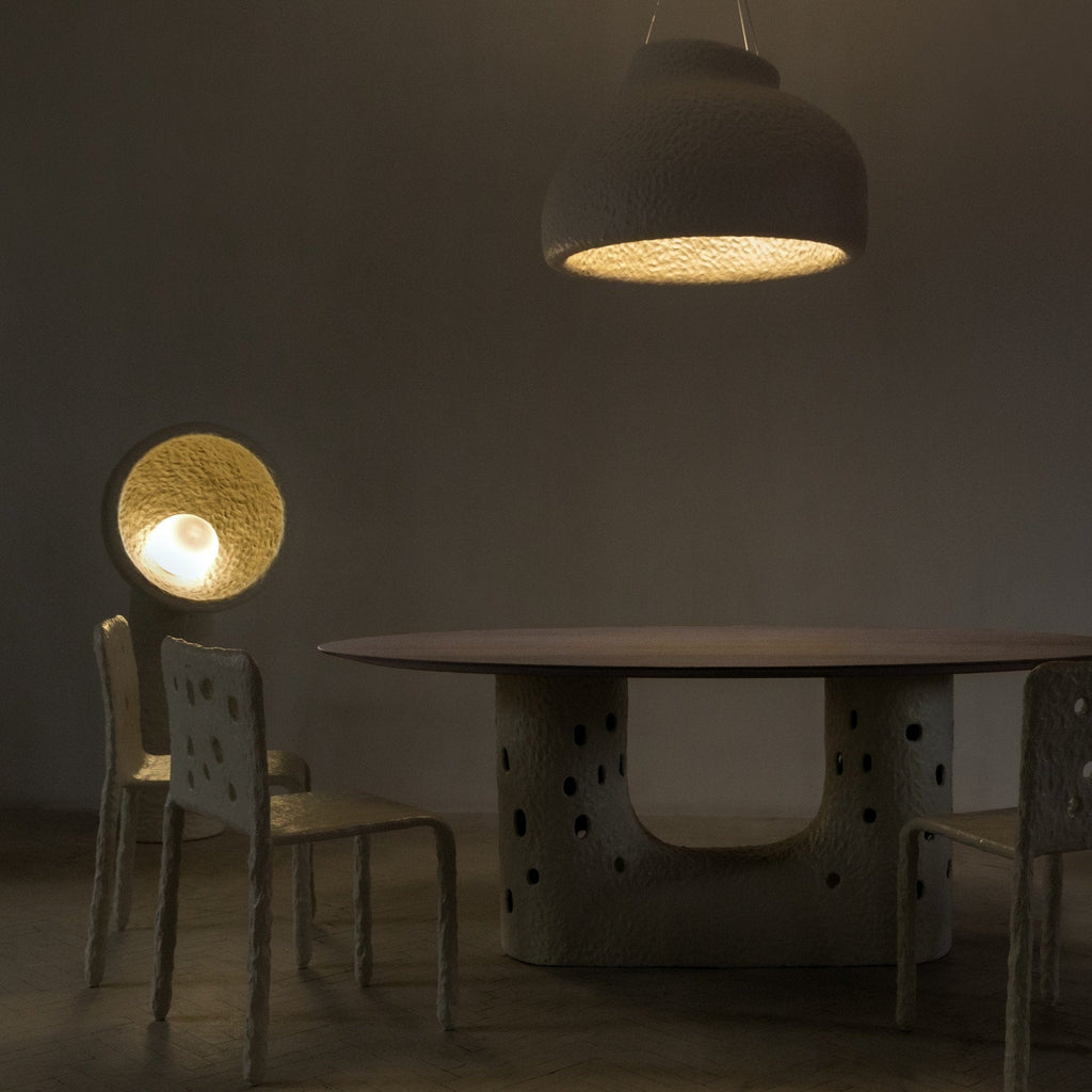 Lit Clérachic floor lamp glowing gold beside a round dark table, with chairs and a hanging dome light overhead.