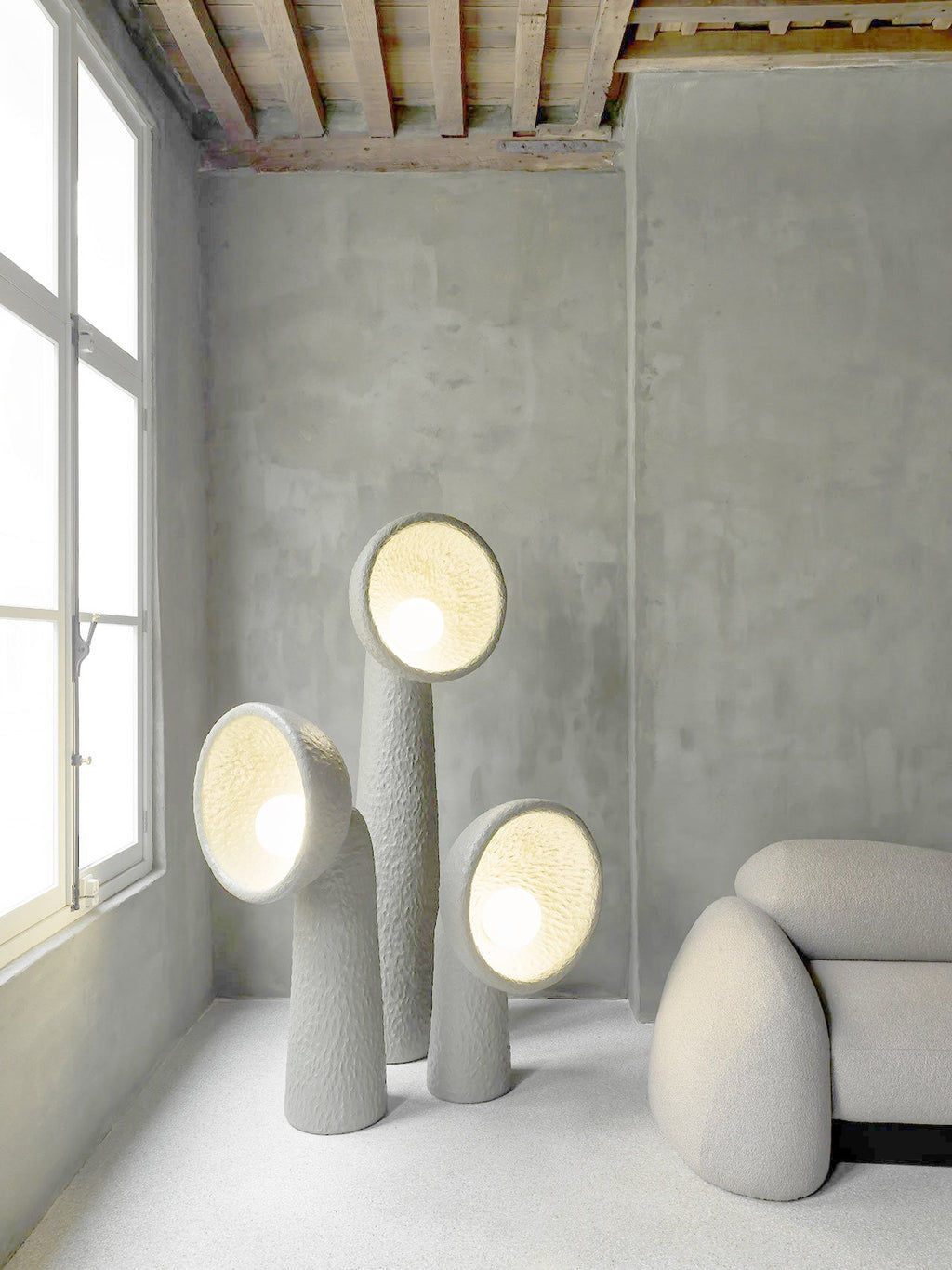 Three Clérachic floor lamps of different heights lit from within, in a grey concrete room beside a tall window.