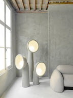 Three Clérachic floor lamps of different heights lit from within, in a grey concrete room beside a tall window.
