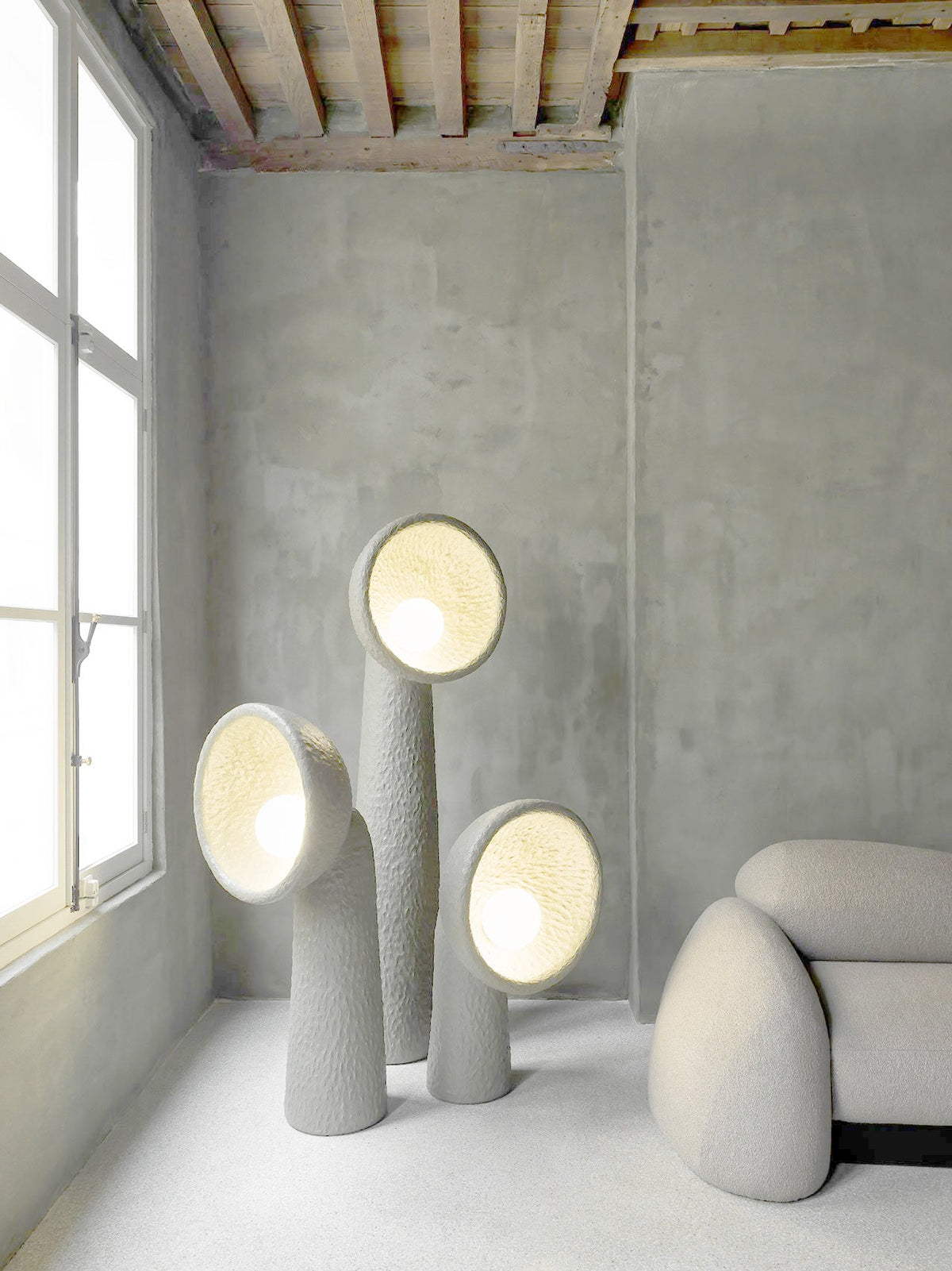 Three Clérachic floor lamps of different heights lit from within, in a grey concrete room beside a tall window.