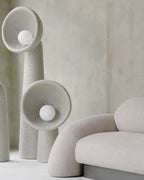 Close view of two white Clérachic floor lamps with bowl shades and round bulbs, beside a pale curved sofa.