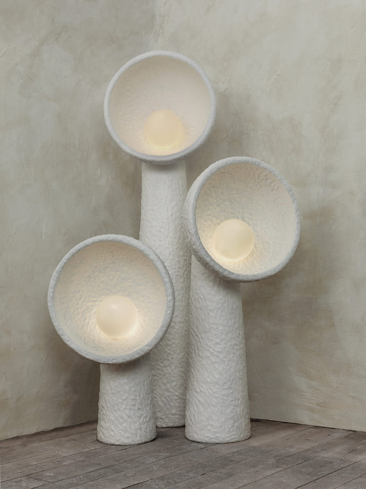 Three Clérachic floor lamps glowing warm in a corner, their rough white bowls lit from inside above a wooden floor.