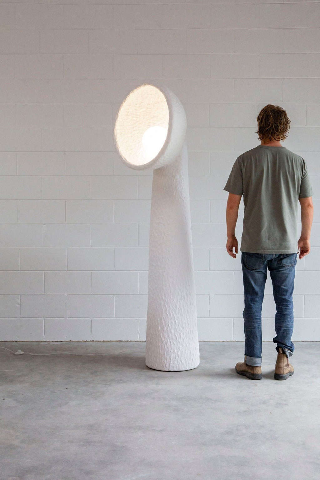 Man standing beside a tall white Clérachic floor lamp in a white-walled room, with a lit bowl shade at head height.