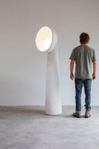 Man standing beside a tall white Clérachic floor lamp in a white-walled room, with a lit bowl shade at head height.