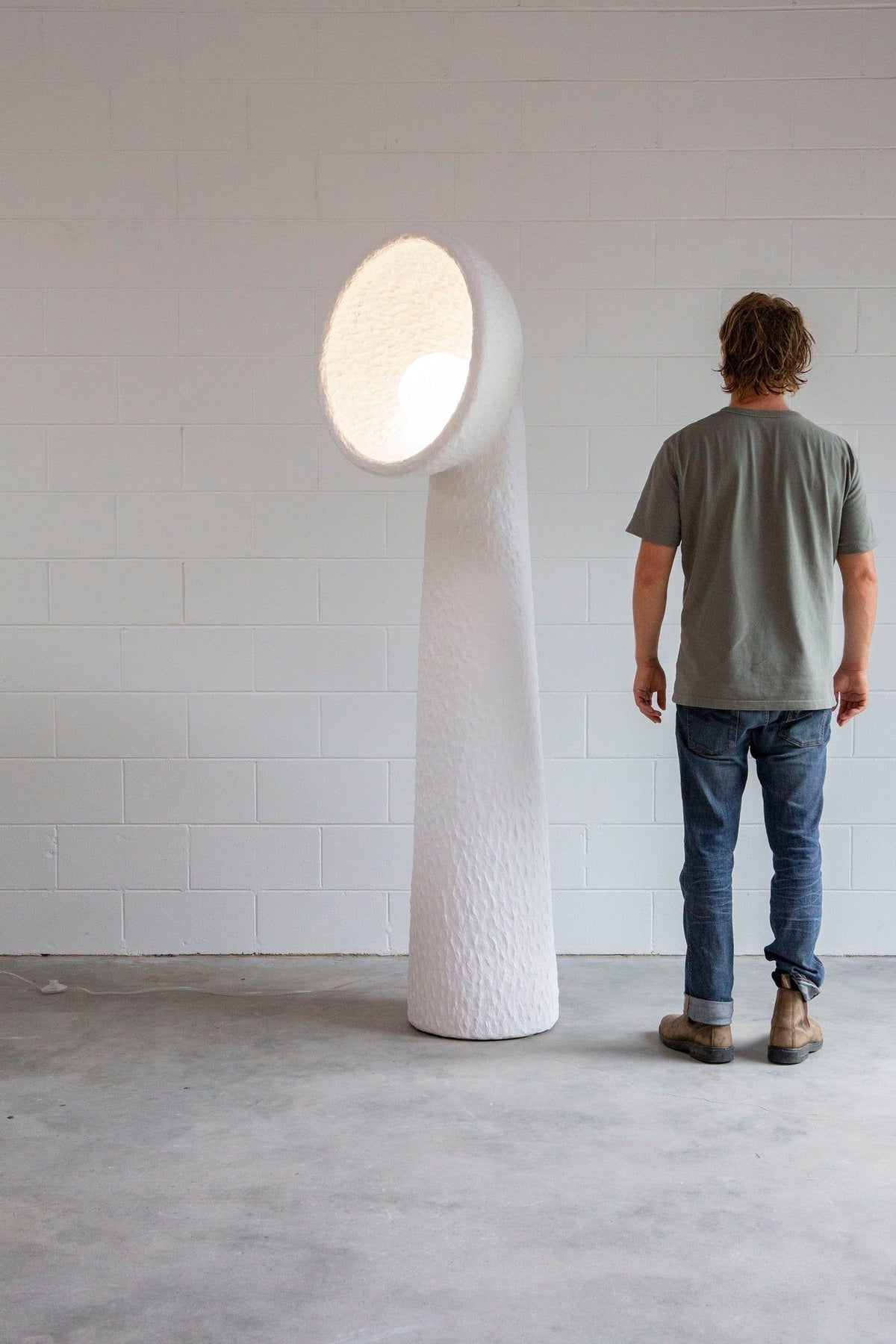 Man standing beside a tall white Clérachic floor lamp in a white-walled room, with a lit bowl shade at head height.
