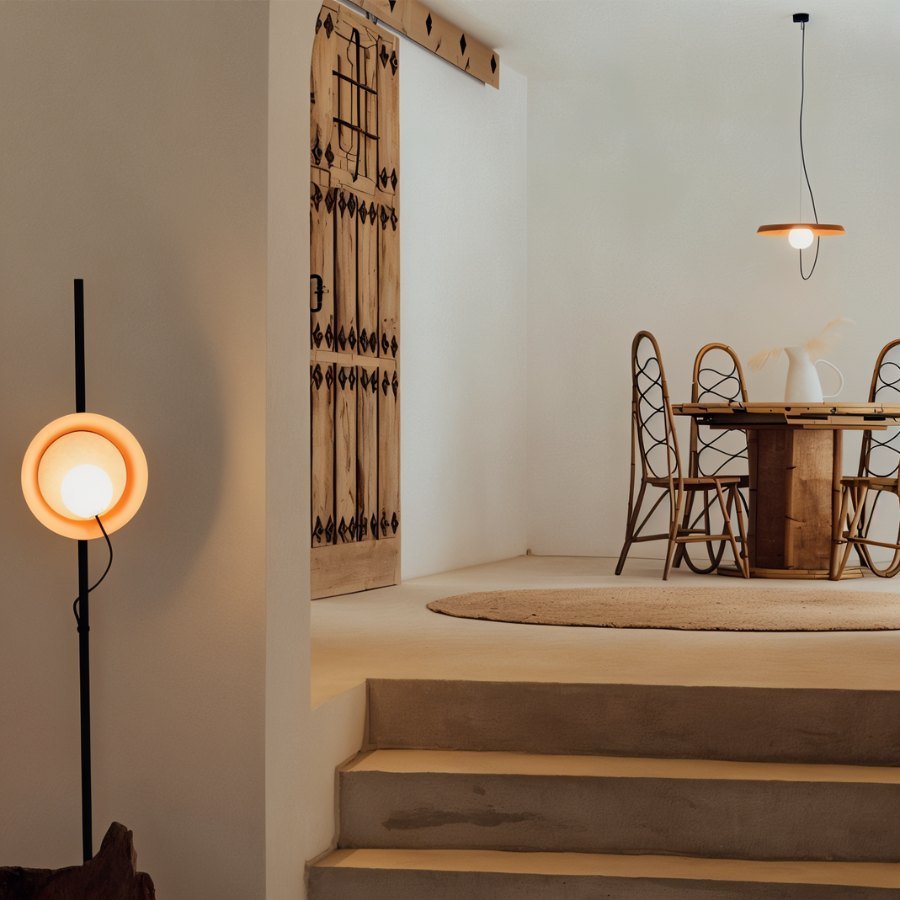 Clérachic disc pendant glowing above a rattan dining set, with a lit floor lamp and carved wooden door beside it.