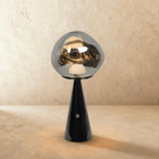 Clérachic table lamp with a chrome-toned distorted glass shade on a glossy black conical base, glowing on a limewash backdrop.