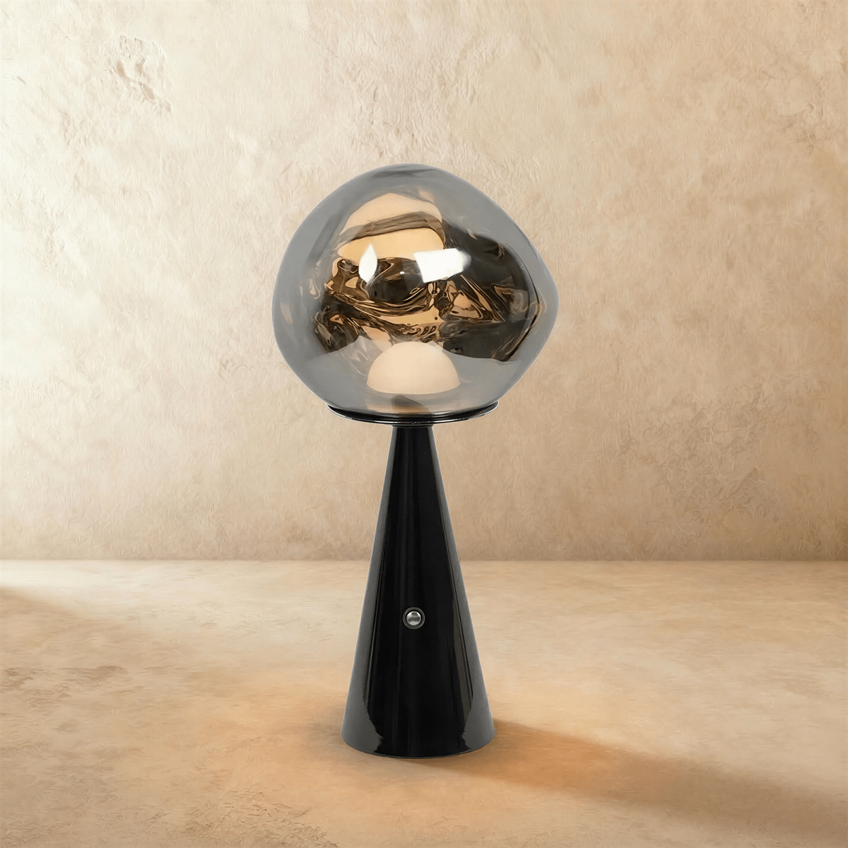 Clérachic table lamp with a chrome-toned distorted glass shade on a glossy black conical base, glowing on a limewash backdrop.