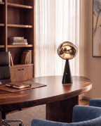 Clérachic table lamp with a glowing distorted glass shade on a black cone base, on a walnut desk beside a leather notebook.
