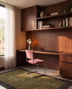 Clérachic chrome-toned table lamp glowing warm on a walnut desk beside a pink velvet chair and a built-in bookcase.