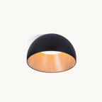 A black Clérachic dome ceiling light with a wood-effect oak interior and a soft white glow at the centre, on a plain off-white backdrop.
