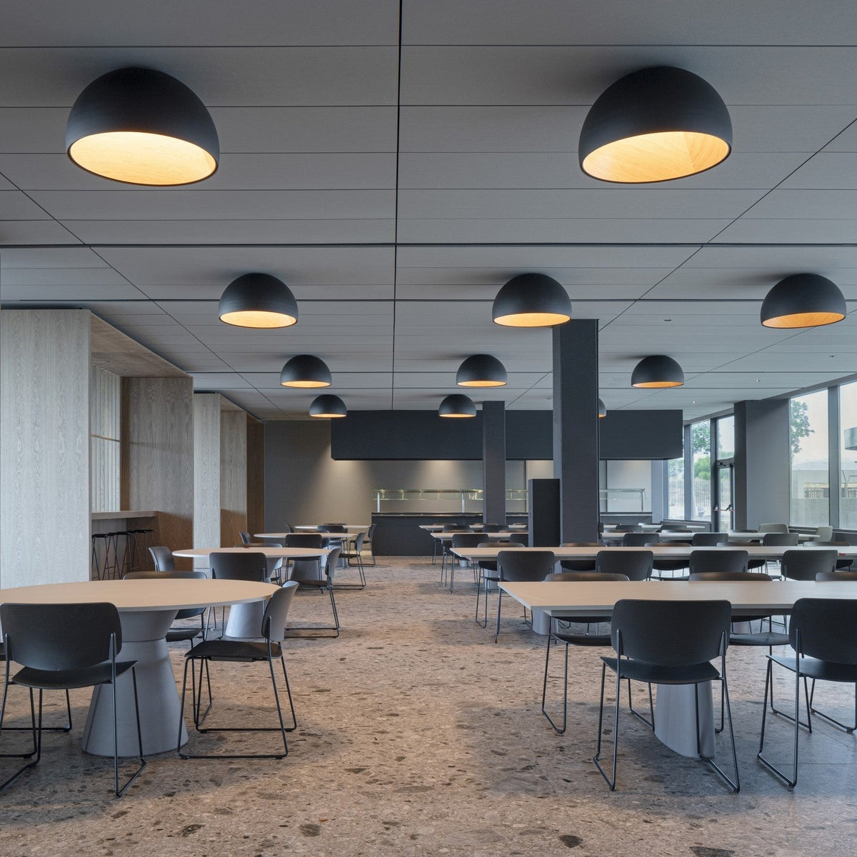 Black dome Clérachic ceiling lights lit at several points across a large canteen with round tables, black chairs and a speckled stone floor.