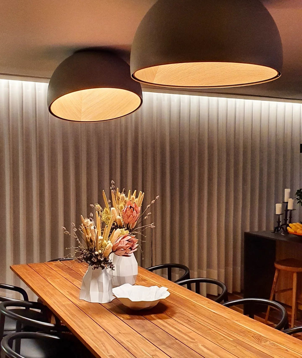 Two black dome Clérachic ceiling lights of different sizes glow over a long timber dining table with dried flowers and a pleated wall.